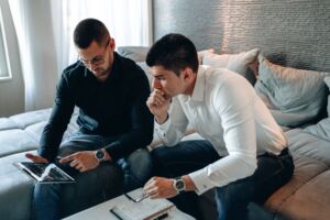 Two men sitting on a couch closely examine a tablet displaying financial charts