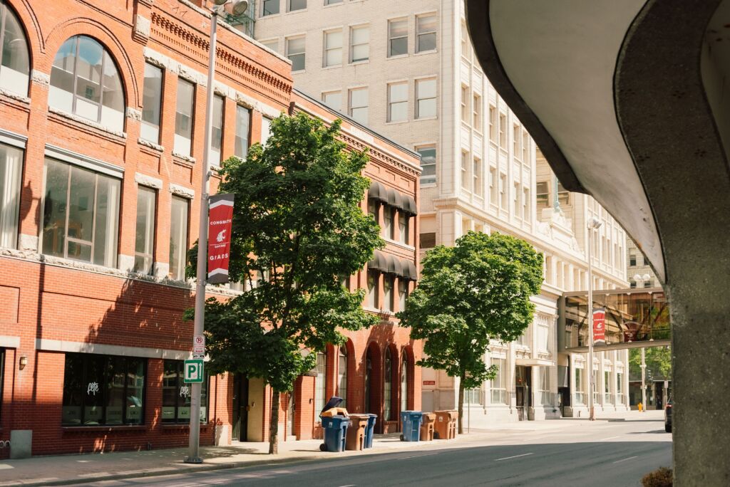 Quiet downtown street with historic buildings, trees, and a skybridge on a sunny day.