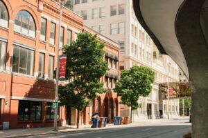 Quiet downtown street with historic buildings, trees, and a skybridge on a sunny day.
